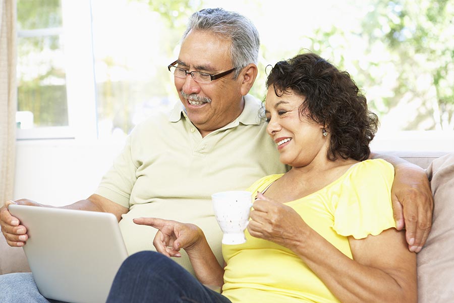 Blog - Senior Couple Smile and Read a Laptop as the Wife Sips Tea, Sitting on Their Sofa on a Sunny Day