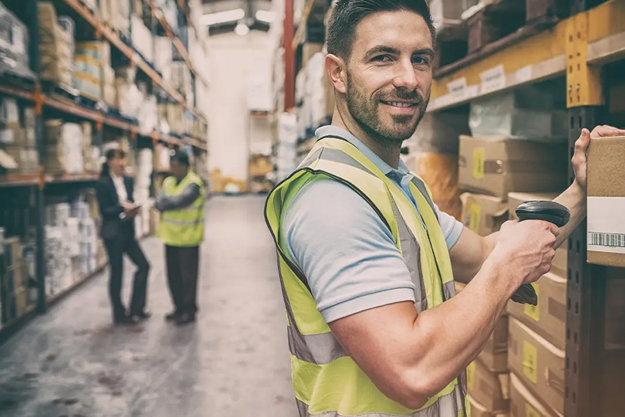 Warehousing and Logistics Insurance - Portrait of a Warehouse Worker Scanning a Box While Smiling With Fellow Colleagues Blurred in the Distance